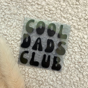 Könnte beinhalten: Ein quadratischer, halbtransparenter Untersetzer aus Kunstharz mit einem dezenten Glitzer-Finish. Die Oberfläche zeigt den schwarzen, fetten und abgerundeten Schriftzug "COOL DADS CLUB" in drei Zeilen. Die Buchstaben wirken glatt und glänzend.