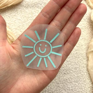 Könnte beinhalten: Ein durchscheinender, kreisförmiger Aufkleber mit einer hellblauen Grafik einer lächelnden Sonne mit strahlenden Strahlen. Das Design besteht aus einem zentralen Kreis mit einer einfachen geschwungenen Linie als Mund und zwei Punkten als Augen.