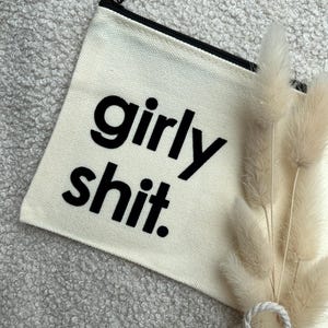 Könnte beinhalten: Eine cremefarbene Reißverschlusstasche aus Segeltuch mit dem schwarzen Schriftzug "girly shit." auf der Vorderseite. Die Tasche hat einen schwarzen Reißverschluss und ist mit einem kleinen Bündel getrockneter, flauschiger Ziergräser verziert.