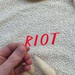 Könnte beinhalten: Ein rechteckiger, transparenter Aufkleber mit dem Wort RIOT in fetten, roten Großbuchstaben. Der Aufkleber hat einen durchsichtigen Hintergrund und wird vor einer strukturierten, cremeweißen Stoffoberfläche mit getrockneten Gräsern gehalten.