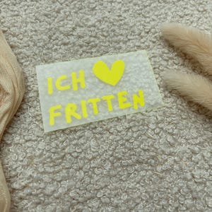 Könnte beinhalten: Ein rechteckiger, halbtransparenter Aufkleber mit dem leuchtend neongelben Text "ICH", gefolgt von einem neongelben Herzsymbol und dem Wort "FRITTEN" in der zweiten Zeile, alles in einer fetten, serifenlosen Schriftart.