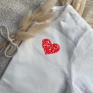 Könnte beinhalten: Ein weißes kurzärmeliges T-Shirt mit einem roten Herzmotiv auf der Brust. Das Herzdesign ist mit einem sich wiederholenden Leopardenmuster gefüllt. Das T-Shirt ist mit einem dekorativen Bündel aus getrocknetem beigem Gras verziert, das mit einer Kordel gebunden ist.