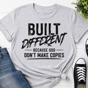 May include: A light gray short-sleeve t-shirt featuring black text that reads BUILT DIFFERENT BECAUSE GOD DON'T MAKE COPIES. The shirt is displayed alongside blue denim jeans and black and white sneakers, with a small green plant visible.