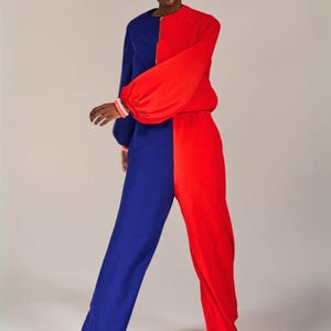 Könnte beinhalten: Ein langärmeliger Jumpsuit mit einem auffälligen Color-Blocking-Design, bei dem die linke Seite in Dunkelblau und die rechte Seite in leuchtendem Rot gehalten ist. Das Kleidungsstück hat einen hohen Kragen mit Reißverschluss und gerade Hosenbeine.