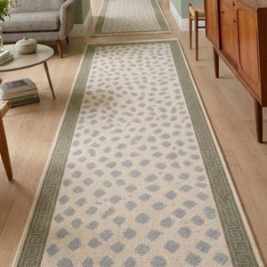 May include: A long rectangular runner rug featuring a cream-colored base with a repeating pattern of soft blue dots. The rug is framed by a wide sage green border with a geometric Greek key design and finished with short fringe on both ends.