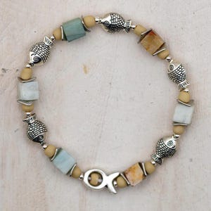 May include: A beaded bracelet featuring silver-toned fish charms, square-shaped stone beads in shades of blue and tan, and small round tan beads. The bracelet is finished with a silver-toned fish-shaped clasp that connects the two ends together.