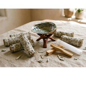 Puede incluir: Una concha de abulón sobre un soporte de madera, rodeada de manojos de salvia blanca, palos de palo santo y una varita de selenita. La concha tiene colores iridiscentes, y los manojos de salvia están atados con hilo. Los artículos están dispuestos sobre una superficie de tono neutro.