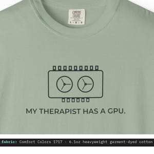 以下が含まれることがあります： セージグリーンのTシャツで、胸の中央にデュアルファン搭載のグラフィックボードの黒い線画が描かれています。グラフィックの下には「MY THERAPIST HAS A GPU.」というテキストがあります。Comfort Colorsブランドのネックラベルが付いています。