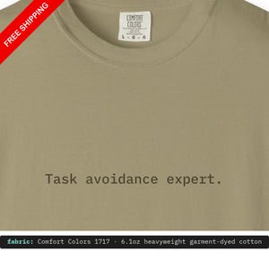 May include: A sage green crew neck t-shirt featuring the text Task avoidance expert. printed in a dark brown typewriter-style font on the chest. The garment includes a white interior neck label with the brand name Comfort Colors.