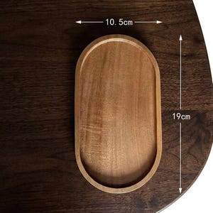 May include: An oval wooden tray with a smooth, light brown finish and a raised rim. The tray features clear measurement markings indicating a width of 10.5 cm and a length of 19 cm, which is approximately 4.13 inches by 7.48 inches in total size.
