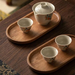 May include: Two oval wooden serving trays holding a ceramic tea set. One tray features a lidded teapot with a built-in strainer and a small cup, both decorated with orange fruit motifs. The second tray holds two matching small ceramic tea cups.