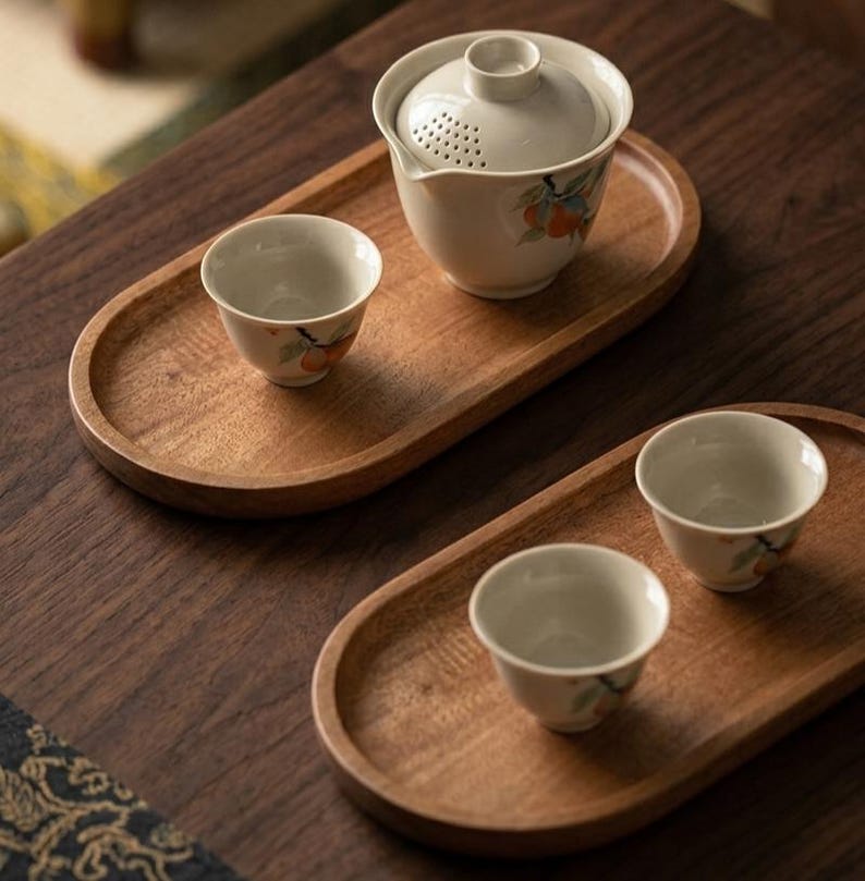 May include: Two oval wooden serving trays holding a ceramic tea set. One tray features a lidded teapot with a built-in strainer and a small cup, both decorated with orange fruit motifs. The second tray holds two matching small ceramic tea cups.