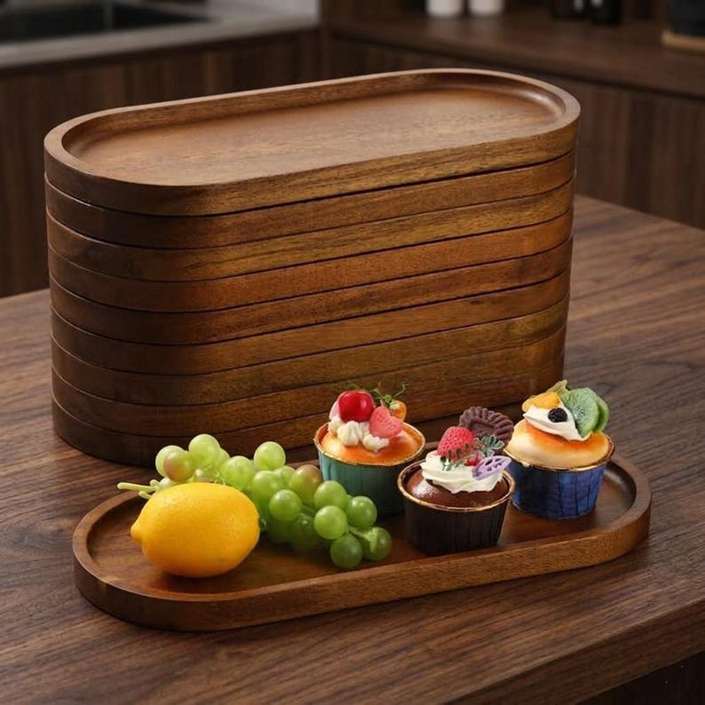 May include: A stack of eight oval wooden trays with a smooth, dark brown finish and raised edges. One identical tray is placed in front of the stack, holding a yellow lemon, a cluster of green grapes, and three cupcakes with various toppings.