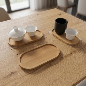 May include: Three oval bamboo trays with raised edges. One tray holds a white lidded bowl and a small white cup. Another tray holds a matte black cup and a small white cup. The third tray is empty. All items are arranged on a wooden surface.