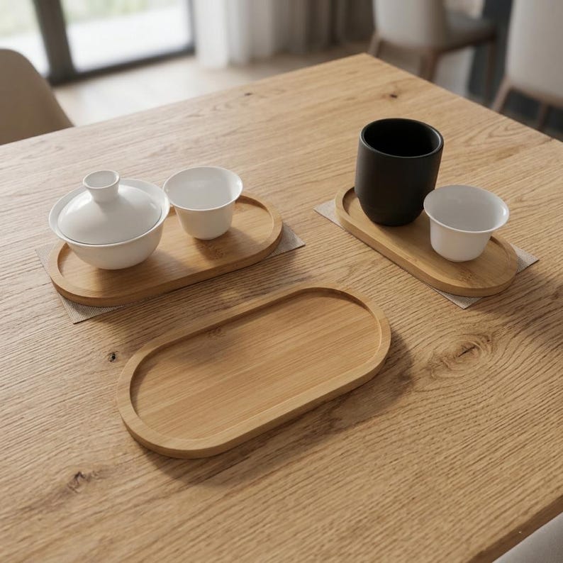May include: Three oval bamboo trays with raised edges. One tray holds a white lidded bowl and a small white cup. Another tray holds a matte black cup and a small white cup. The third tray is empty. All items are arranged on a wooden surface.