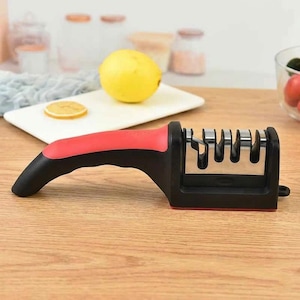 Op de afbeelding: Een handmatige messenslijper met een ergonomisch zwart en rood handvat en een metalen slijpkop met drie afzonderlijke sleuven voor het slijpen van messen. Het apparaat heeft een stevige basis en een compact ontwerp voor gebruik in de keuken.