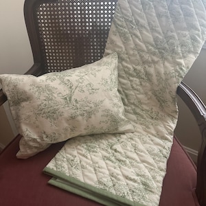 Peut inclure: Un oreiller décoratif et une couverture matelassée avec un motif de toile de Jouy vert et blanc. L'oreiller et la couverture sont posés sur une chaise bordeaux avec un dossier en cannage. La couverture est drapée sur la chaise.