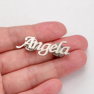 Peut inclure: Une épingle de couleur argentée avec le nom "Angela" écrit en cursive. L'épingle est tenue dans une main, et le nom est fixé à deux attaches.