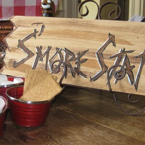 May include: A wooden sign with the words "S'more Station" painted in white on a brown background. The sign is propped up on a metal stand. There are four red metal buckets filled with marshmallows and graham crackers.