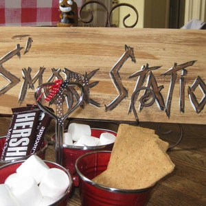 May include: A wooden sign with the words "S'more Station" painted in white on a brown background. There are three red bowls filled with marshmallows, a chocolate bar, and graham crackers.