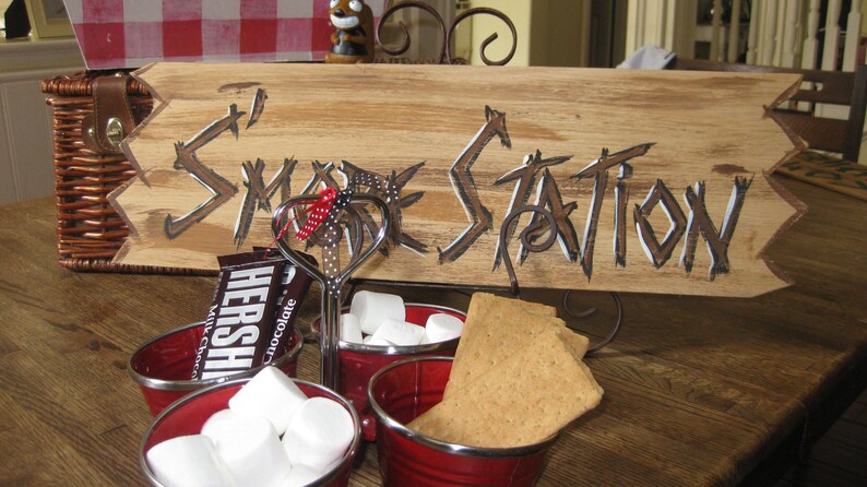 May include: A wooden sign with the words "S'more Station" painted in white on a brown background. There are three red bowls filled with marshmallows, a chocolate bar, and graham crackers.