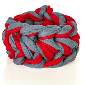 Könnte beinhalten: Geflochtenes Armband in Rot und Grau. Das Armband besteht aus dicken, gewebten Stoffstreifen, die einen klobigen, strukturierten Look erzeugen. Die roten und grauen Farben sind gleichmäßig verteilt und schaffen ein auffälliges Accessoire.