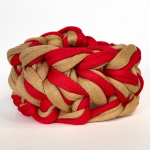 May include: A braided fabric bracelet with alternating tan and red stripes. The bracelet is made of a soft, woven material and has a unique, textured design.