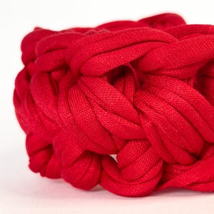 May include: A close-up of a red knitted bracelet made from thick yarn. The bracelet is woven in a complex pattern, creating a textured and intricate design.