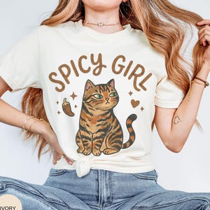 May include: An ivory short-sleeve t-shirt featuring a brown graphic of a tabby cat sitting upright. Above the cat, the text reads SPICY GIRL in brown letters. A small brown chili pepper and a heart icon are positioned on either side of the cat.