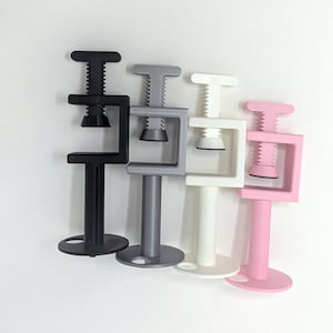 May include: Four small, colorful clamps in black, gray, white, and pink. Each clamp has a T-shaped handle, a threaded rod, and a C-shaped frame. The clamps are arranged in a row, with the bases of the clamps resting on a white surface.