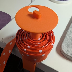 May include: An orange ribbon spool with white polka dots. The spool is held by an orange plastic holder. The ribbon is rolled up and ready to use. A gray felt mat is in the background.