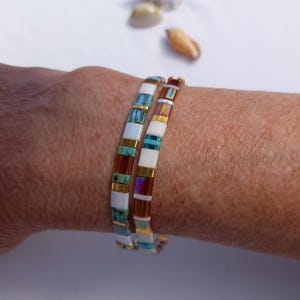 Op de afbeelding: Twee gestapelde armbanden gemaakt van rechthoekige kralen in een herhalend patroon van wit, turkoois, bruin en iriserend metallic goud. De kralen zijn in een lineaire volgorde gerangschikt, wat een kleurrijk en gestructureerd ontwerp op de pols creëert.