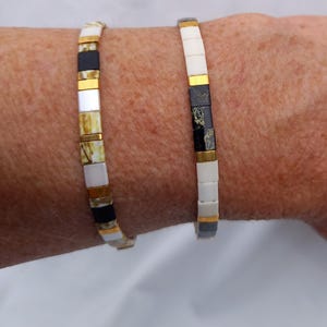 Op de afbeelding: Twee kralenarmbanden met een herhalend patroon van vierkante tegeltjes in wit, matzwart en metallic goud. De linkerarmband bevat gemarmerde beige en witte tegeltjes, terwijl de rechterarmband effen witte en zwart gespikkelde tegeltjes toont.