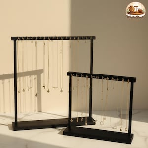 May include: Two black wooden jewelry display stands. The stands are rectangular with a series of small pegs along the top for hanging necklaces. Several gold necklaces are displayed on the stands. The stands are on a white surface.