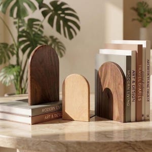 May include: Three arch-shaped wooden bookends with smooth surfaces. Two bookends are made of dark brown walnut wood, and one smaller bookend is made of light-colored oak wood. They are positioned to support a stack of hardcover books.