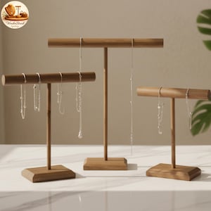May include: Three wooden jewelry display stands in varying heights. Each stand has a T-shaped design with a rectangular base and a horizontal bar for hanging necklaces. The stands are made of light brown wood and are holding silver necklaces.