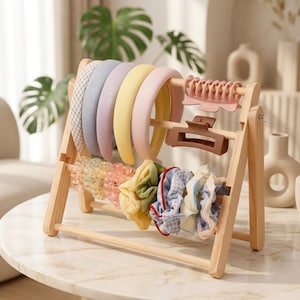 May include: A light-colored wooden A-frame display stand holding five fabric-covered headbands in various pastel colors, two plastic hair claws in pink and brown, and a row of assorted fabric scrunchies in yellow, green, and blue patterns.