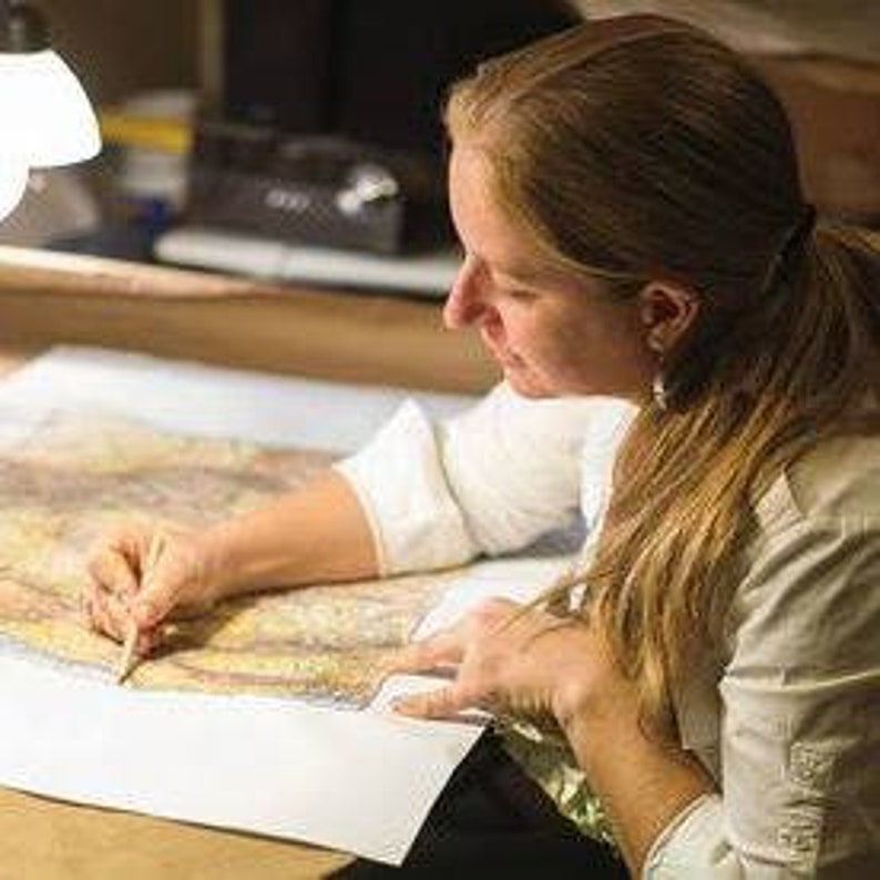 K&ouml;nnte beinhalten: Eine Frau arbeitet mit einem Bleistift an einer gro&szlig;en Karte. Die Karte liegt auf einem Tisch ausgebreitet und zeigt ein detailliertes geografisches Gebiet.