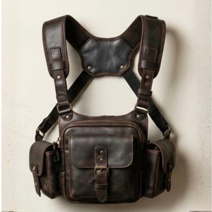 May include: A brown leather chest bag with multiple pockets and adjustable straps. The bag features a central compartment with a buckle closure and two side pockets. The design includes a padded shoulder and back support for comfort.