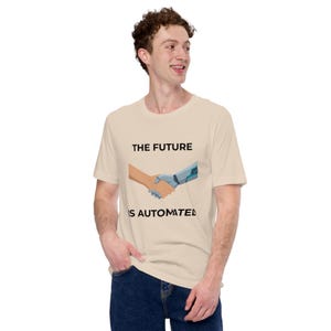 May include: A light beige t-shirt with the text "THE FUTURE IS AUTOMATED" above an illustration of a human hand shaking a robotic hand. The shirt is worn by a person.