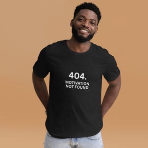 Op de afbeelding: Zwart T-shirt met de witte tekst "404. MOTIVATION NOT FOUND". Het T-shirt heeft een ronde hals en lijkt gemaakt van een zacht materiaal. De persoon die het T-shirt draagt, glimlacht.