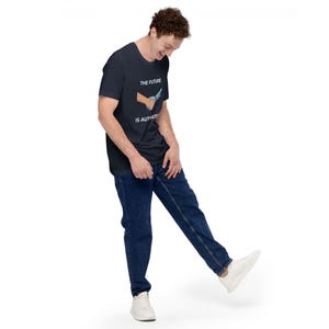May include: Navy blue t-shirt with the text "THE FUTURE IS AUTHENTIC" and a graphic of a handshake with a knife. The shirt is paired with dark blue jeans and white sneakers. The person is smiling and standing on a white background.