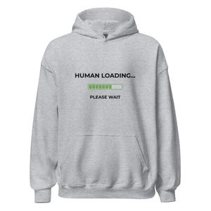 Könnte beinhalten: Hellgrauer Kapuzenpullover mit Kapuze und Vordertasche. Der Text "HUMAN LOADING..." steht über einem grünen Ladebalken, darunter "PLEASE WAIT". Ein lässiges, bequemes Kleidungsstück.