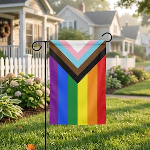 May include: A garden flag featuring the Progress Pride flag design. The flag has a black pole and is displayed in a grassy yard with houses in the background. The flag's design includes the traditional rainbow colors, along with pink, light blue, brown, black, and white.