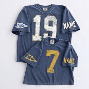 May include: Two blue short-sleeve t-shirts with distressed graphics. The larger shirt features the number 19 in white, a pennant with the text MASCOT, and the word NAME. The smaller shirt features the number 7 in yellow, a yellow pennant with MASCOT, and NAME.