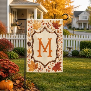 May include: A rectangular garden flag featuring a cream-coloured centre with a large orange letter M. The border is decorated with brown branches, orange and red autumn maple leaves, pinecones, and small berries, all suspended on a black metal stand.