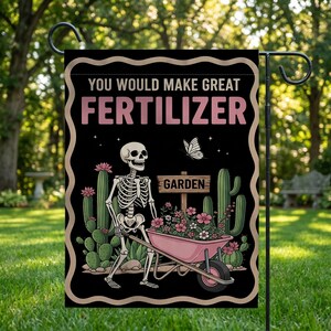 Könnte beinhalten: Schwarze Gartenflagge mit humorvollem Design. Sie zeigt ein Skelett, das eine rosa Schubkarre voller Blumen schiebt. Der Text lautet "YOU WOULD MAKE GREAT FERTILIZER" oben und "GARDEN" auf einem Schild. Kakteen und ein Schmetterling sind ebenfalls abgebildet.