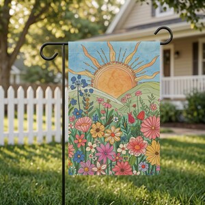 May include: A garden flag featuring a colorful illustration of a sunrise over a field of wildflowers. The design includes a large orange sun, green hills, and a variety of flowers in shades of pink, yellow, blue, and red. The flag is displayed outdoors.