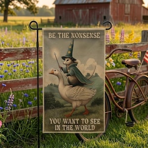 Puede incluir: Bandera de jardín con el texto "BE THE NONSENSE YOU WANT TO SEE IN THE WORLD" y una ilustración de un mago zarigüeya montando un ganso. La bandera está ambientada en un entorno rural con un granero rojo y una bicicleta vintage.