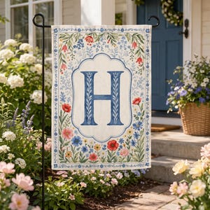 May include: A cream-colored garden flag featuring a large blue letter "H" in the center, surrounded by a floral border with red, pink, and blue flowers. The flag is displayed on a black metal stand, perfect for outdoor decor.